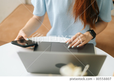 Close-up front view of unrecognizable young woman in smart watch using laptop and phone sitting at table in cafe with warm daylight, selective focus. 80558807