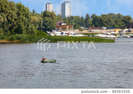 A fisherman on an inflatable rubber boat is fishing with fishing rods in the middle of the river on a sunny summer day. 80563508
