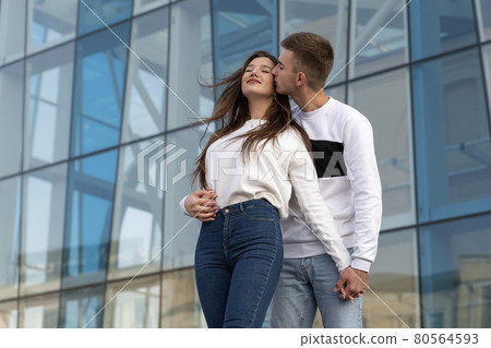 Guy hugs and kisses his girlfriend on the street. Young couple in love. City building on the background Guy hugs and kisses his girlfriend on the street. Young couple in love. City building on the background 80564593
