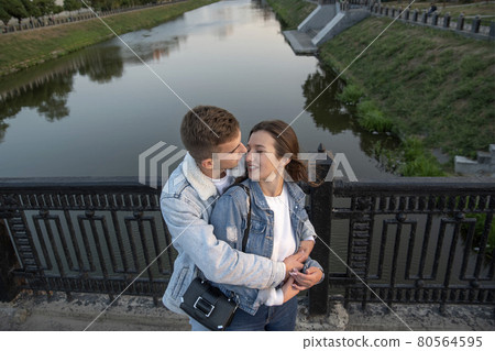 Happy couple embraces on the bridge. Guy hugs and kisses his beloved on river background. Above view. Romantic date Happy couple embraces on the bridge. Guy hugs and kisses his beloved on river background. Above view. Romantic date 80564595