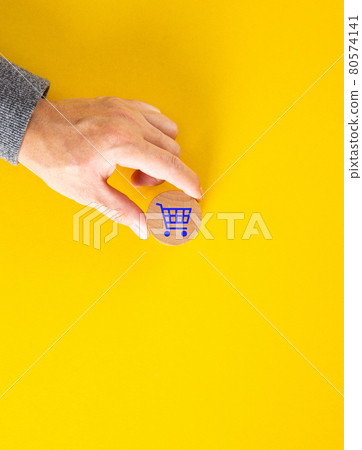 Male hand placing a blue shopping cart icon on yellow background. Male hand placing a blue shopping cart icon on yellow background. 80574141