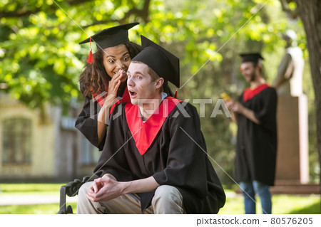 A cute girl in academic cap whispering something to her friend in the ear 80576205