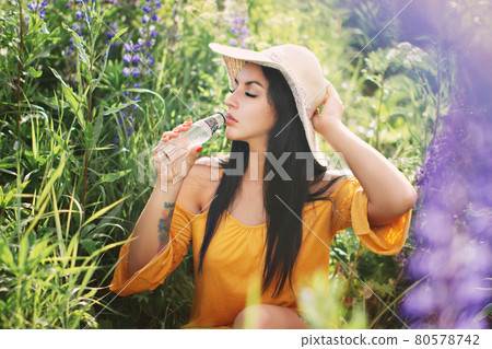 Woman in straw hat and yellow dress drinks water from glass bottle in field of lupins in summer 80578742
