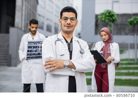 Arabic or hindu man doctor in front of multiethnic team, looking at camera with arms crossed Arabic or hindu man doctor in front of multiethnic team, looking at camera with arms crossed 80586710