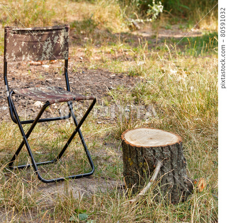 A canvas folding tourist chair sits next to a makeshift table in the form of a wooden stump. A canvas folding tourist chair sits next to a makeshift table in the form of a wooden stump. 80591032