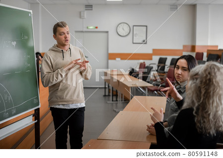 Two girls and a guy are talking in sign language. Three deaf students chatting in a university classroom. Two girls and a guy are talking in sign language. Three deaf students chatting in a university classroom. 80591148