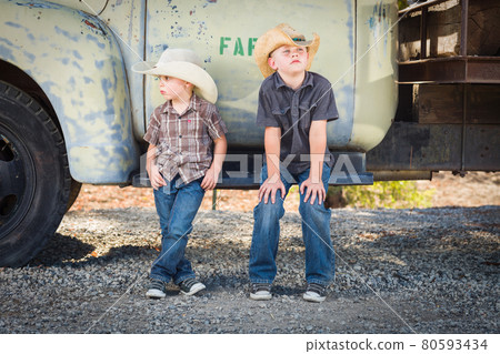 Two Young Boys Wearing Cowboy Hats Leaning Against an Antique Truck in a Rustic Country Setting.. 80593434