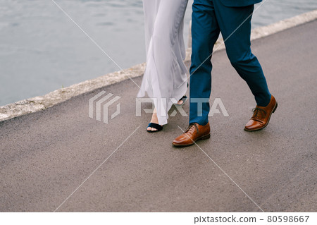 Legs of the groom in blue trousers and the bride in a white dress are walking along the asphalt road over the sea. Close-up 80598667