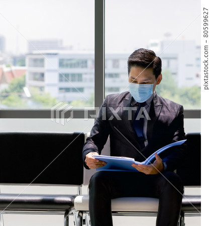 Asian business man in black suit and face mask sit on a bench near the window, examines his resume while waiting for the interview. Asian business man in black suit and face mask sit on a bench near the window, examines his resume while waiting for the interview. 80599627