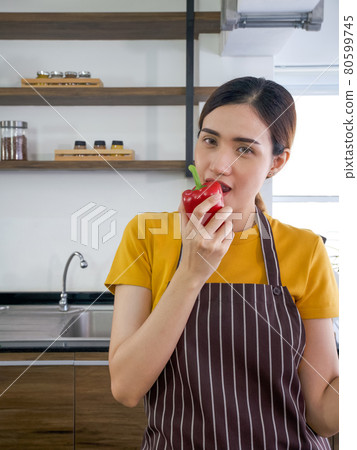 The housewife dressed in an apron bites the delicious red bell pepper. Close up The housewife dressed in an apron bites the delicious red bell pepper. Close up 80599745