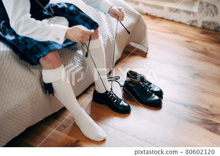 Preparing for a Scottish wedding. Man in a kilt, sporran and high socks sits on the bed and tie long shoelaces on his shoes Preparing for a Scottish wedding. Man in a kilt, sporran and high socks sits on the bed and tie long shoelaces on his shoes 80602120