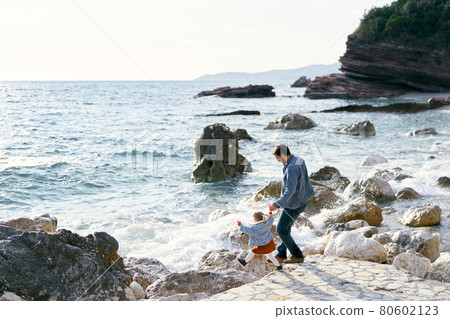 Dad holds the hand of a little girl, going down with her along a stone path to a pebble beach to the water against a background of mountains. Back view 80602123