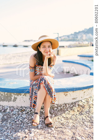 Smiling girl in a hat and dress sits on a bench on a pebble beach Smiling girl in a hat and dress sits on a bench on a pebble beach 80602201