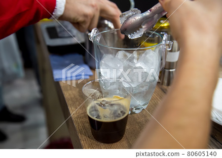 a hand with tongs takes ice from a vase to put in a plastic glass with black freshly brewed coffee. close-up, soft focus, blur background 80605140