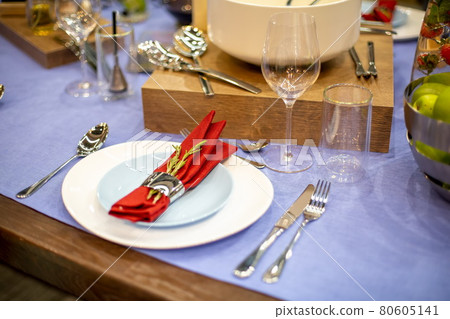 table setting with a blue tablecloth, a white and blue plate, a red napkin in a steel holder with a branch of rosemary, steel appliances, glass goblets. close up, soft focus 80605141