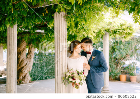 Smiling groom hugs the bride's shoulders against the background of columns in a green garden Smiling groom hugs the bride's shoulders against the background of columns in a green garden 80605522