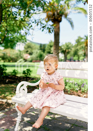 Little barefoot girl in a dress sits on a bench with a leaf in her hand Little barefoot girl in a dress sits on a bench with a leaf in her hand 80605532