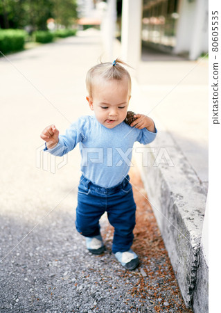 Little girl with a ponytail walks along the road in the park and carries a pebble on her shoulder 80605535