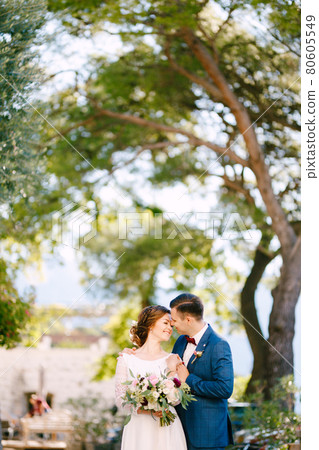 Groom gently hugs the bride by the shoulders against the backdrop of trees on a warm sunny day. Bride holds a beautiful bouquet of flowers in her hands Groom gently hugs the bride by the shoulders against the backdrop of trees on a warm sunny day. Bride holds a beautiful bouquet of flowers in her hands 80605549