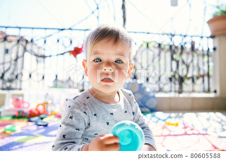Cute baby sitting on a colored rug on the balcony and holding a plastic toy in his hand 80605588