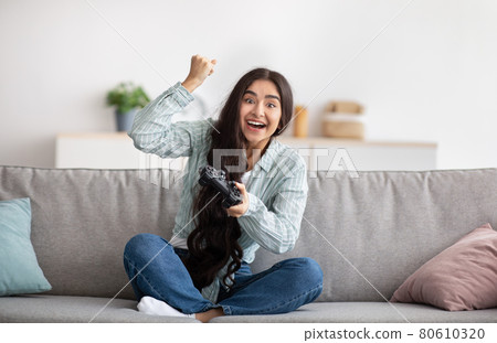 Full length fo excited Indian woman with joystick sitting on couch at home, making victory gesture, winning video game 80610320