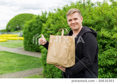 Man with eco paper bag for takeaway food showing thumbs up on nature green background. Delivery in any weather around the clock to the client. 80616363