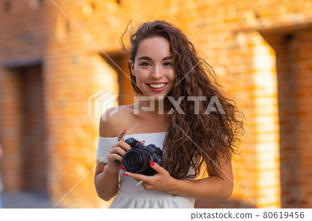 Young attractive female student or tourist using a mirrorless camera while walking in summer city. Woman takes pictures and enjoy weather. 80619456