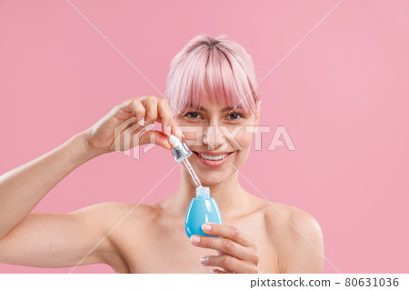 Portrait of smiling young woman with pink hair and bare shoulders looking at camera, holding bottle of serum, posing isolated over pink background 80631036