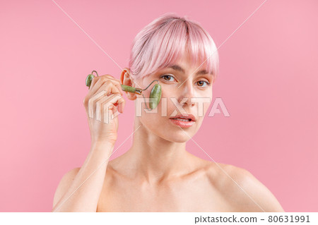 Portrait of young naked woman with pink hair using natural jade facial roller for skin care, posing isolated over pink background Portrait of young naked woman with pink hair using natural jade facial roller for skin care, posing isolated over pink background 80631991