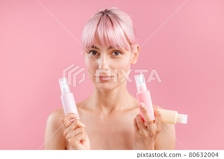 Attractive young woman with pink hair looking at camera, holding three plastic travel bottles with beauty products, posing isolated over pink background Attractive young woman with pink hair looking at camera, holding three plastic travel bottles with beauty products, posing isolated over pink background 80632004