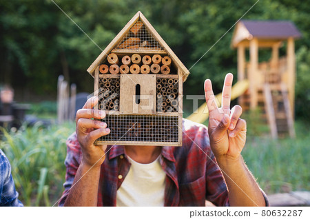 Happy young man with bug hotel spending time outdoors in backyard, showing v sign with fingers. 80632287