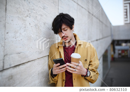 Portrait of young man standing against concrete wall outdoors, using smartphone. 80632310