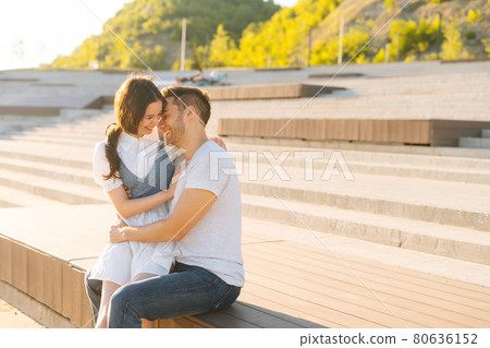 Portrait of cheerful young couple resting in city park sitting higging on bench in summer sunny day. 80636152