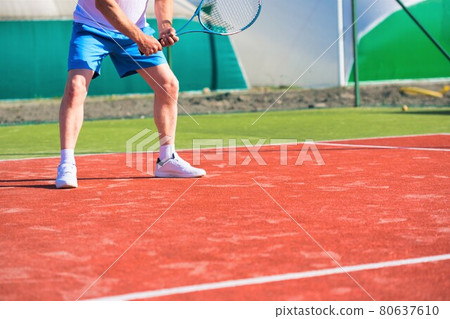 Confident mature man hitting tennis ball with racket on red court 80637610
