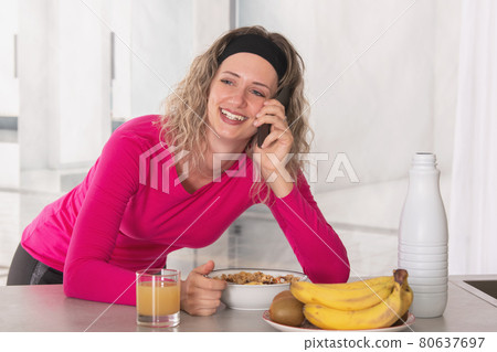 Cheerful woman eating cereal with fruit and milk for breakfast while talking on the phone. Cheerful woman eating cereal with fruit and milk for breakfast while talking on the phone. 80637697