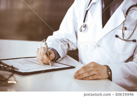 Unknown male doctor sitting and working with clipboard of medication history record in clinic at his working place, close-up. Young physician at work. Perfect medical service, medicine concept 80639946