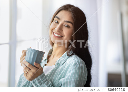 Portrait of pretty Indian lady standing near window with cup of hot drink, smiling at camera, indoors Portrait of pretty Indian lady standing near window with cup of hot drink, smiling at camera, indoors 80640218