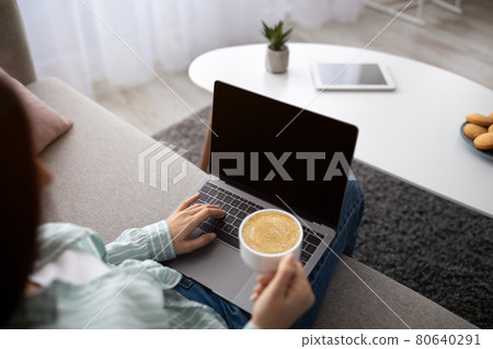 Young Indian woman with coffee sitting on sofa and using laptop computer with blank screen at home, mockup Young Indian woman with coffee sitting on sofa and using laptop computer with blank screen at home, mockup 80640291