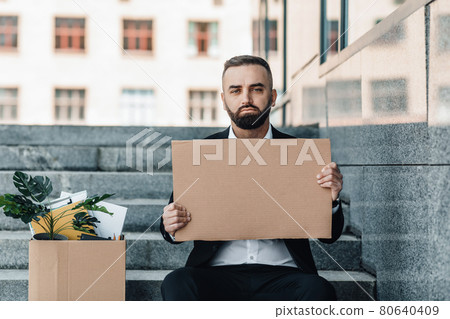 Sad middle-aged businessman sitting with empty poster and box of personal belongings on stairs near office center Sad middle-aged businessman sitting with empty poster and box of personal belongings on stairs near office center 80640409