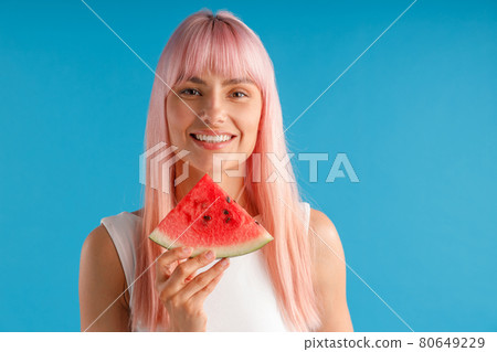Portrait of beautiful young woman with pink hair holding watermelon slice and smiling at camera isolated over blue studio background 80649229