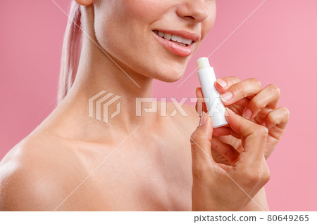 Cropped portrait of smiling woman with naked shoulders holding lip balm near her lips isolated over pink background Cropped portrait of smiling woman with naked shoulders holding lip balm near her lips isolated over pink background 80649265