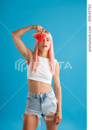 Cute young woman with pink hair holding a slice of watermelon over her eye, standing isolated over blue studio background 80649762