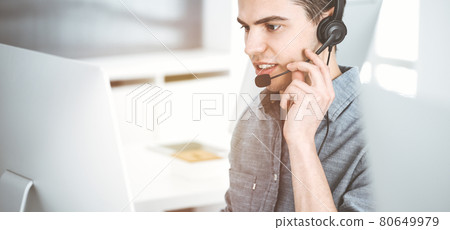 Casual dressed young man using headset and computer while talking with customers online in sunny office. Call center, business concept Casual dressed young man using headset and computer while talking with customers online in sunny office. Call center, business concept 80649979