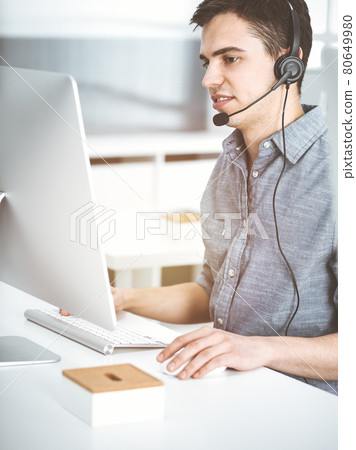 Casual dressed young man using headset and computer while talking with customers online in sunny office. Call center, business concept Casual dressed young man using headset and computer while talking with customers online in sunny office. Call center, business concept 80649980