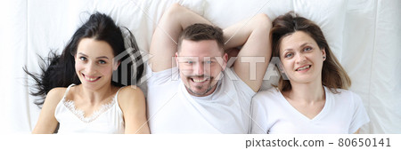 Two smiling women and a man lie on bed 80650141