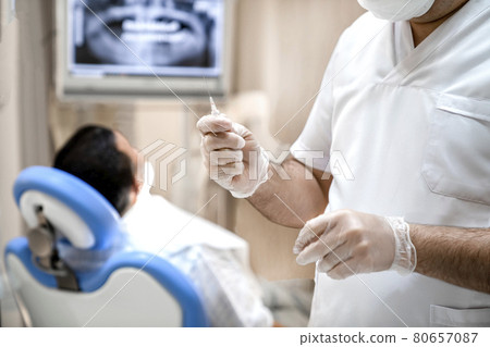 The dentist holds a syringe in his hands with a drug for anesthesia. The patient is waiting for anesthesia while sitting in the dentist's chair. The dentist holds a syringe in his hands with a drug for anesthesia. The patient is waiting for anesthesia while sitting in the dentist's chair. 80657087