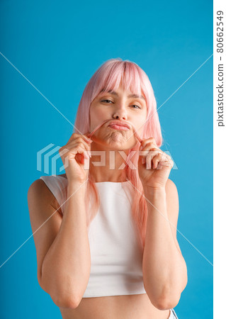 Playful young woman with natural long pink dyed hair holding a strand of it as a moustache, posing isolated over blue studio background 80662549