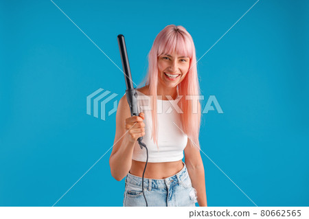 Joyful young woman with pink hair in casual wear smiling at camera, holding curling iron, standing isolated over blue studio background Joyful young woman with pink hair in casual wear smiling at camera, holding curling iron, standing isolated over blue studio background 80662565