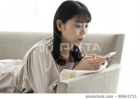 一個女人躺在靠窗的沙發上，正在操作智能手機 80663715