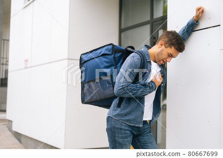 Exhausted food delivery man with large thermo backpack stopping by wall and holding heart, standing in city street. Exhausted food delivery man with large thermo backpack stopping by wall and holding heart, standing in city street. 80667699